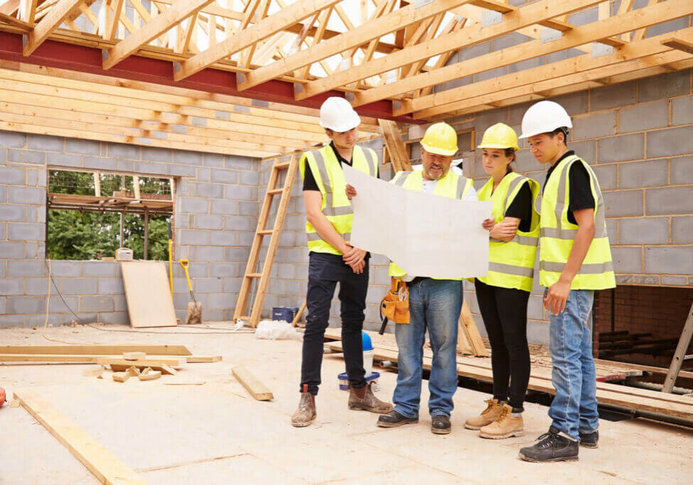 Builder On Building Site Looking At Plans With Apprentices Builder On Building Site Looking At Plans With Apprentices